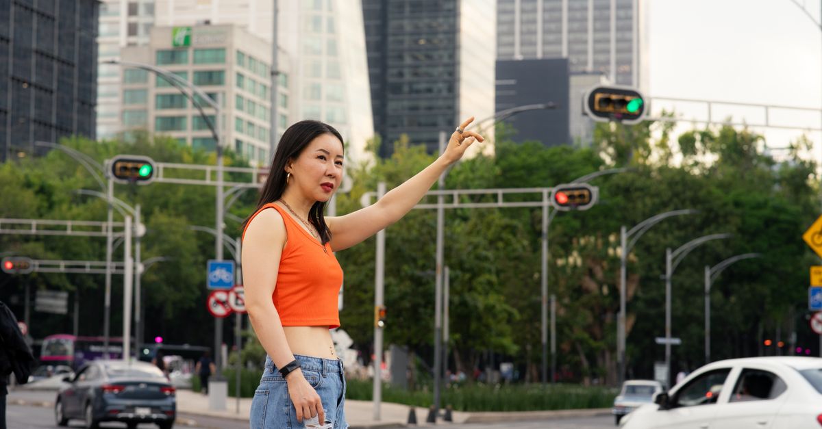 Woman in an orange top and jeans hailing a taxi on a busy city street with skyscrapers.
